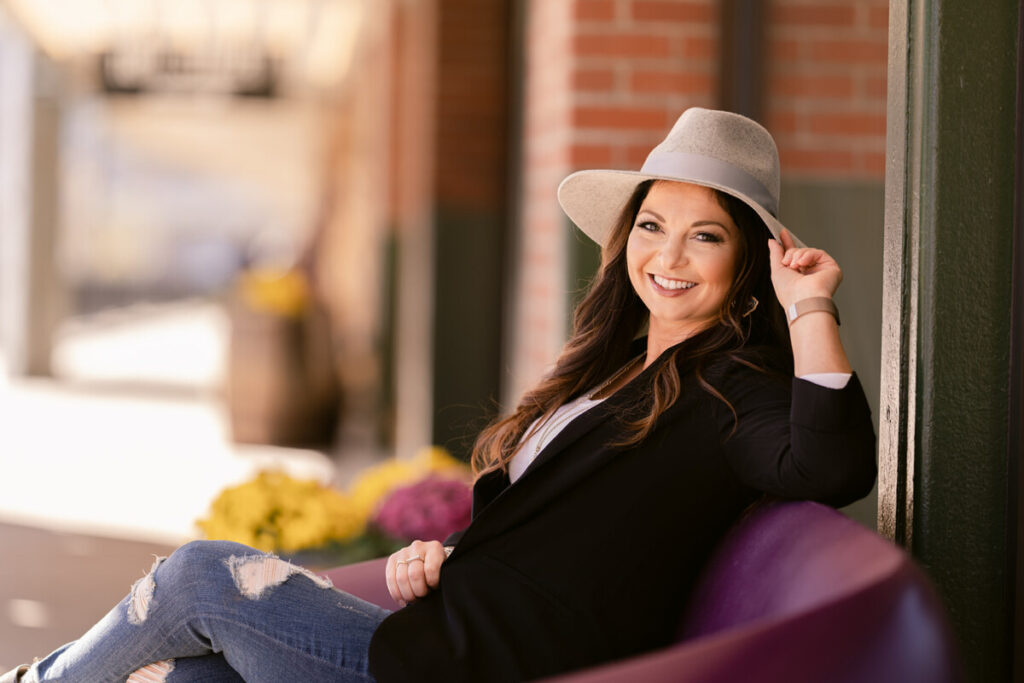 Smiling woman in a stylish hat and blazer, seated outdoors and radiating confidence, symbolizing the journey from manifesting stress to success.