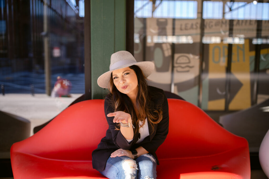 Smiling woman in a hat blowing a playful kiss, sitting on a red couch in a casual outdoor setting. This image connects with the concept of the gut-brain connection, stress, and digestive health, symbolizing the impact of stress relief on overall wellness and joy.