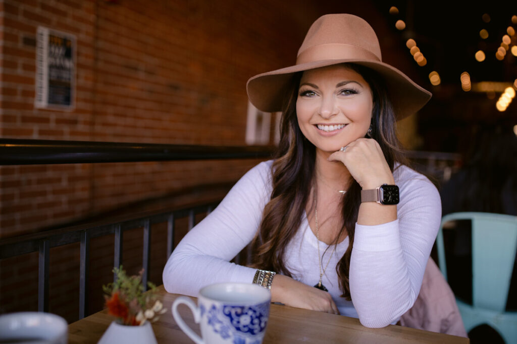 Smiling woman wearing a brown hat, seated in a cozy cafe setting, representing the confidence and calm that comes from mastering the subconscious mind for success.